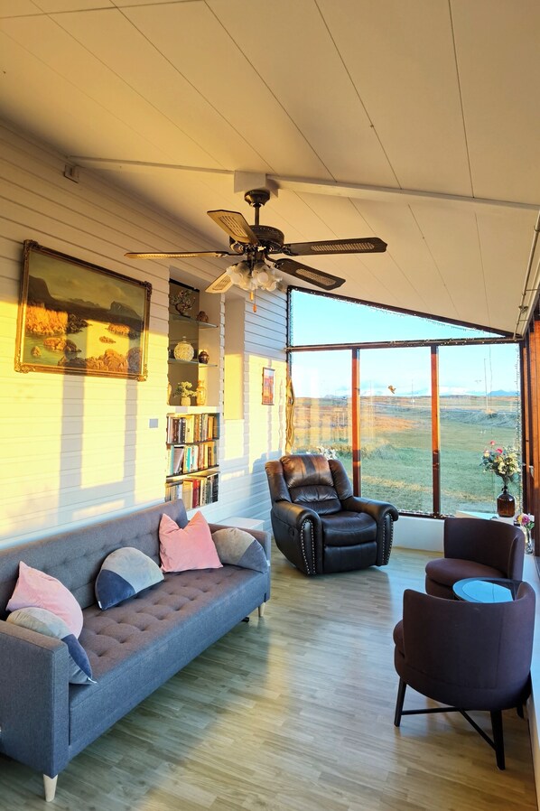 A spacious living room with a high ceiling, a ceiling fan, a large window offering a view of the outdoors, a dark leather armchair, a grey sofa with cushions, and a bookshelf filled with books and decorative items.