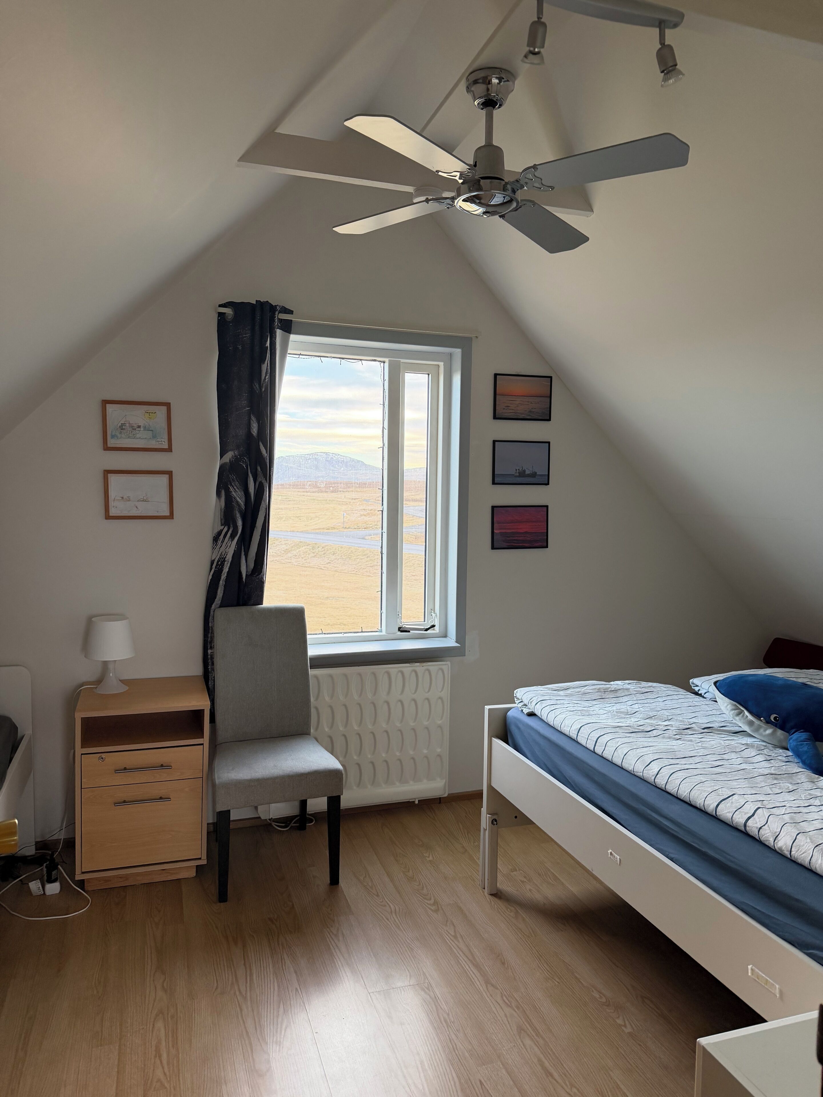 Un dormitorio con cama, escritorio, silla, radiador, ventilador de techo y ventana con vistas a un paisaje desértico.