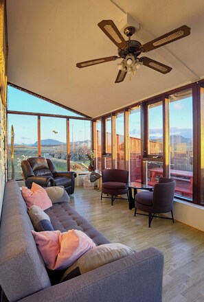 A spacious living room with a large sofa, two armchairs, and a dining table. A ceiling fan is visible, and the room has large windows offering a view of the outdoors.