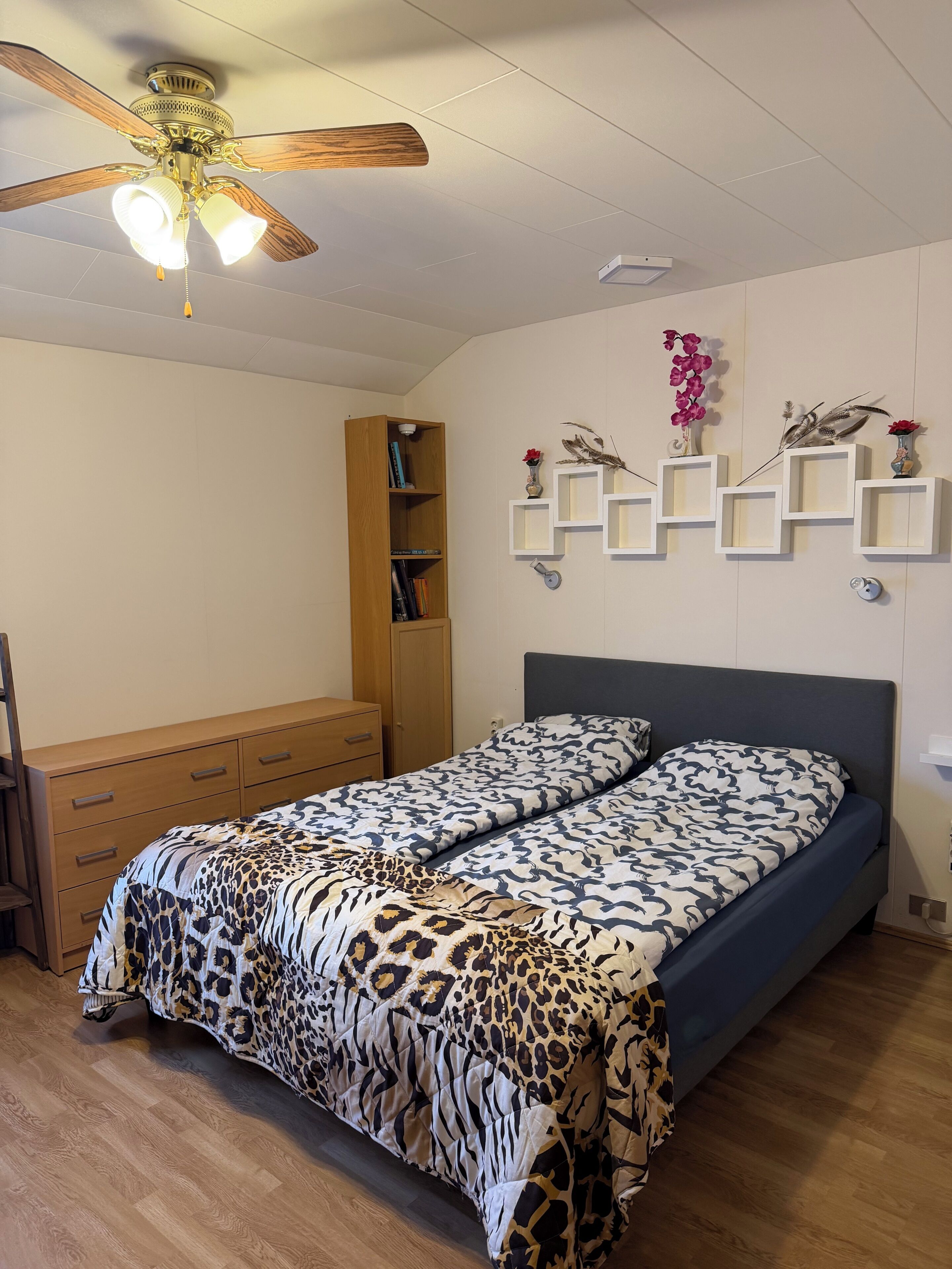 A bedroom with a bed, a wooden wardrobe, a ceiling fan, and wall-mounted shelves with decorative items.