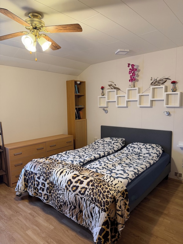 A bedroom with a bed, a wooden wardrobe, a ceiling fan, and wall-mounted shelves with decorative items.