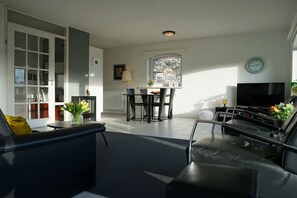 A modern living room with a sofa, a coffee table, a TV, and a dining area with chairs and a table. - Residence Juliana 15 (Julianadorp)