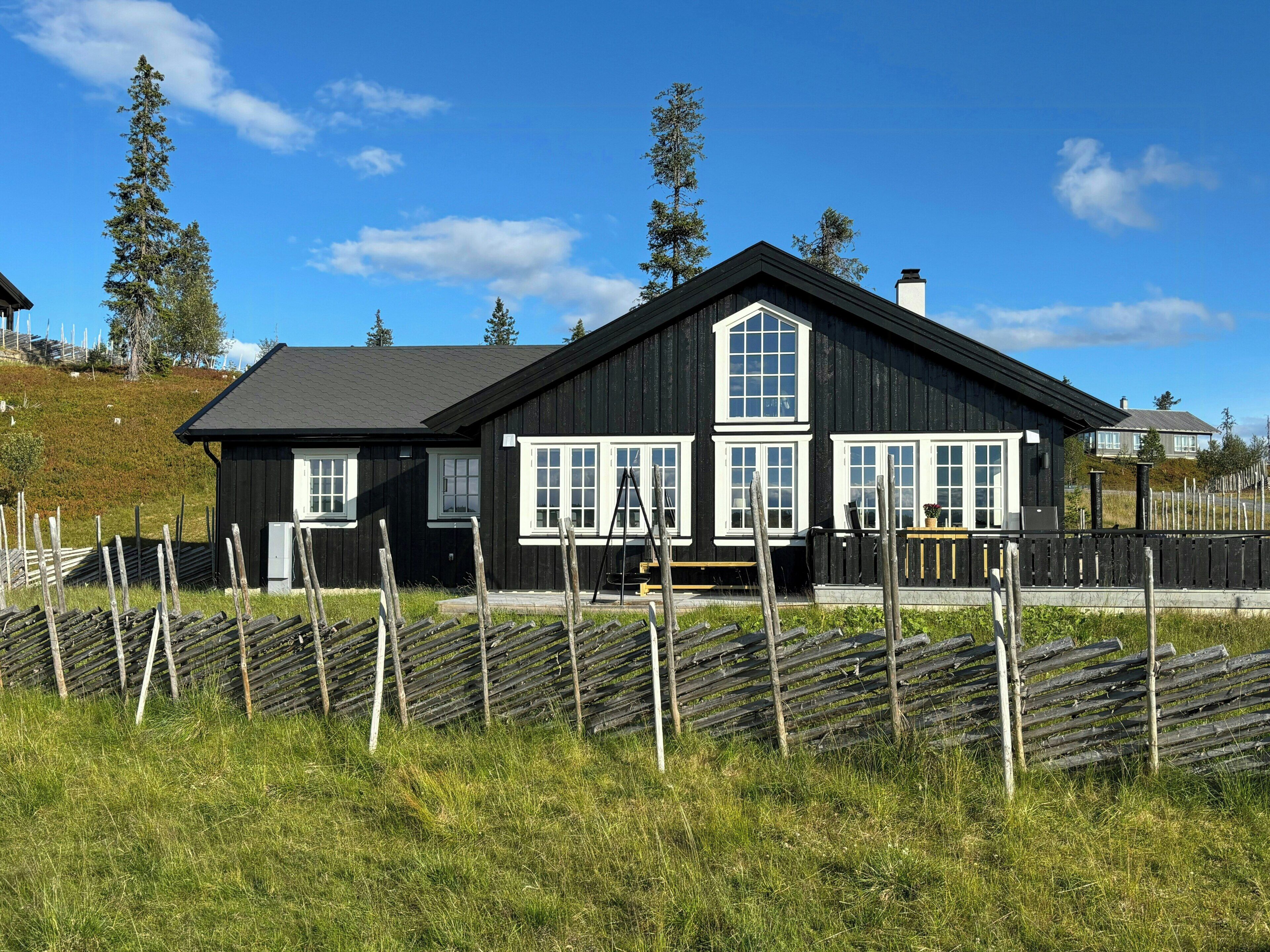 Een zwart houten huis met witte ramen en een schoorsteen, omgeven door een houten hek en gras.