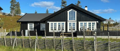 Et sort træhus med hvide vinduer og skorsten, omgivet af et træhegn og græs.