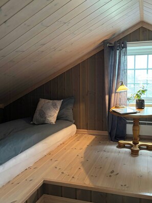 Um quarto de sótão aconchegante, com teto inclinado, cama com almofadas estampadas, mesa com abajur e vista pela janela.