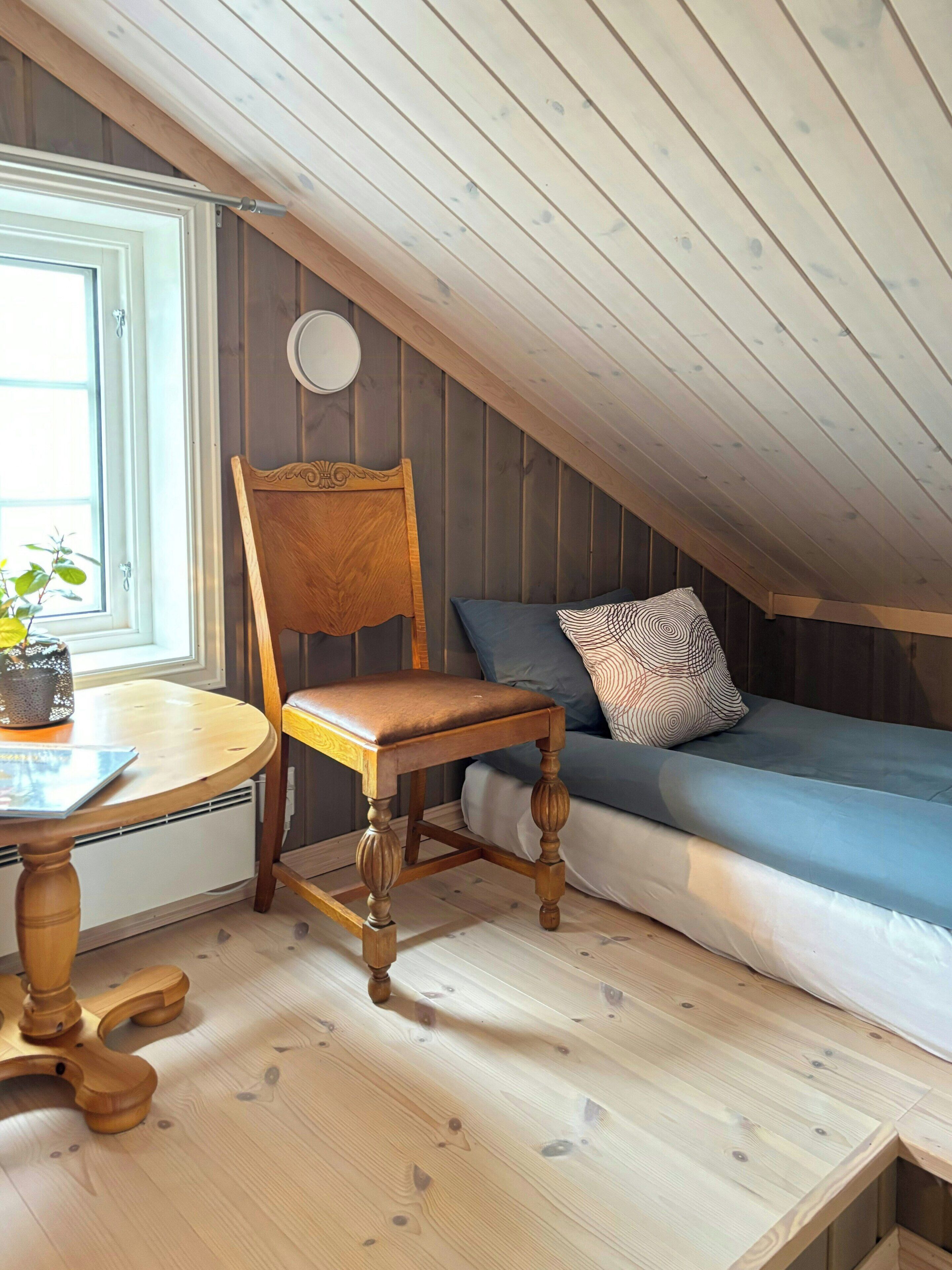 A room with a wooden chair, a small table, a sofa with patterned cushions, and a window with a plant on the table.