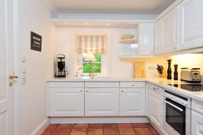 A kitchen with white cabinetry, a coffee machine, a window with a striped blind, and a view of greenery outside. - House on the Cliff - Weberhof (Keitum)