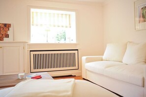 A room with a white radiator, a white sofa, a window with blinds, and a framed picture on the wall. - House on the Cliff - Weberhof (Keitum)