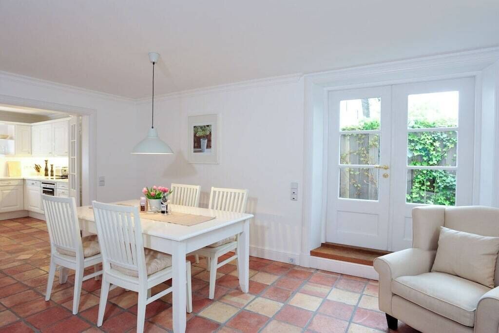 Uma sala de jantar com uma mesa e cadeiras brancas, uma luminária pendente e vista para o jardim através de uma porta.