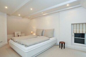 A bedroom with a bed, bedside table, lamp, and a window with blinds. - House on the Cliff - Weberhof (Keitum)