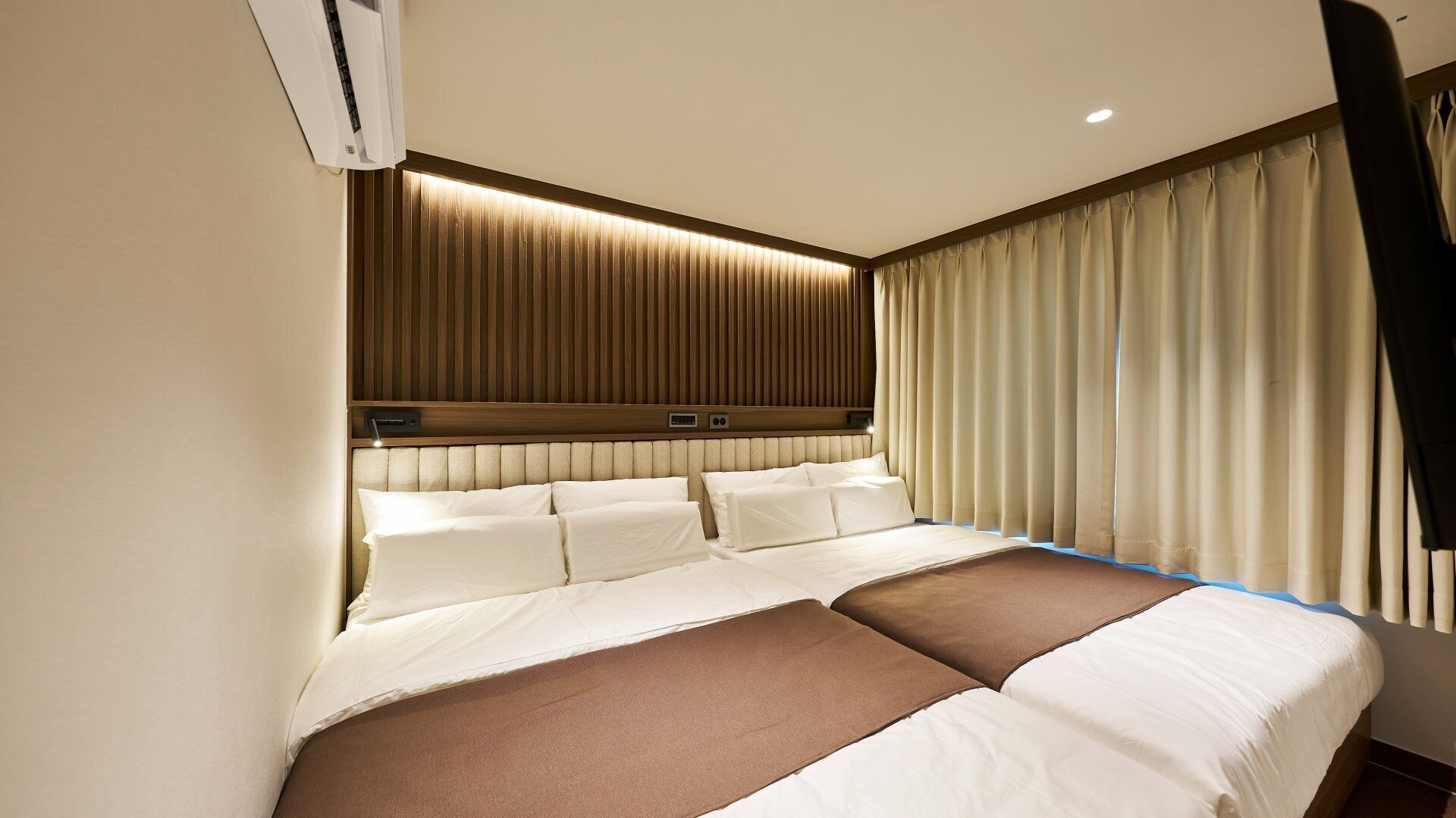 A hotel room with a large bed, a headboard with built-in lighting, and a window with curtains.