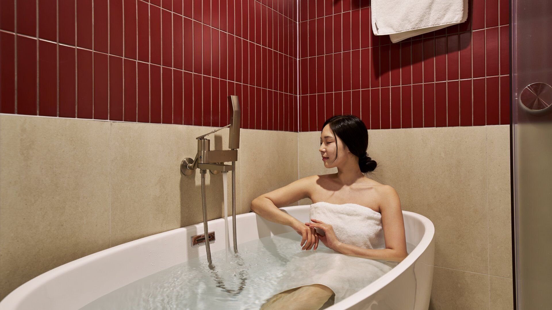 A person in a bathtub with a showerhead, beige walls, and a red tiled background.