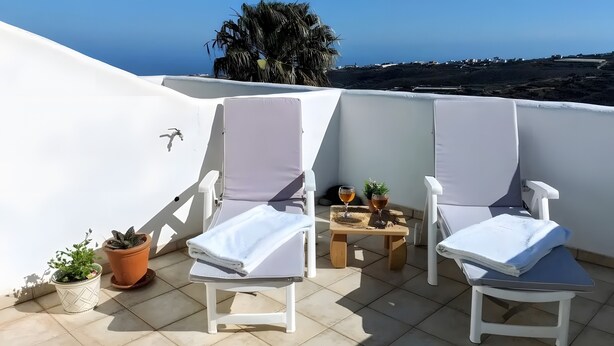 Eine Terrasse mit zwei weißen Liegestühlen, einem kleinen Tisch mit einem Weinglas und Topfpflanzen.