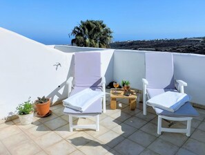 Dues cadires relaxants blanques amb tovalloles, una taula petita amb un got de vi i plantes en testos en una terrassa enrajolada amb vista al mar i una palmera.
