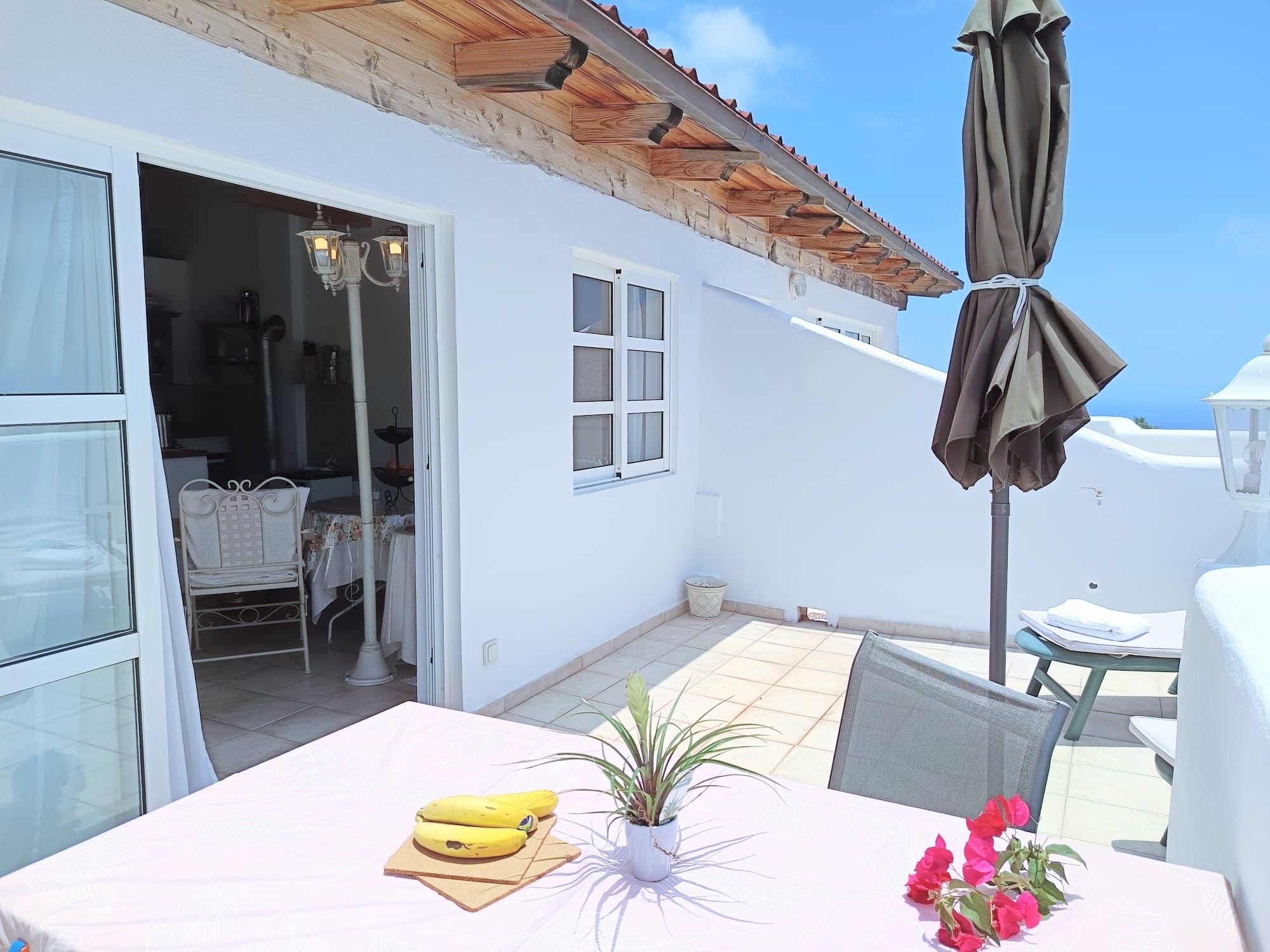 A white building with a tiled patio, a table with a plant and flowers, and a chair.