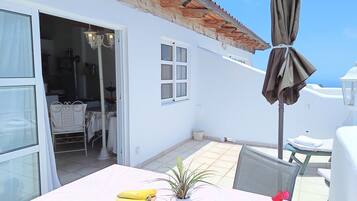 A white building with a tiled patio, a table with a plant and flowers, and a chair.