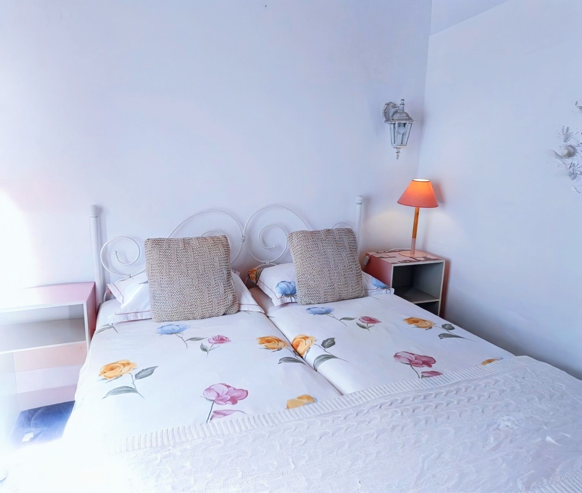 Een bed met een bloemenprint deken, twee kussens, een nachtkastje met een lamp en een wandlamp.