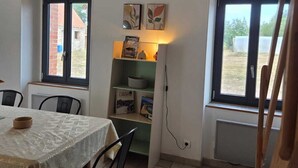 A dining area with a table set for two, a bookshelf with books and decorative items, and a window with a view of the outside.