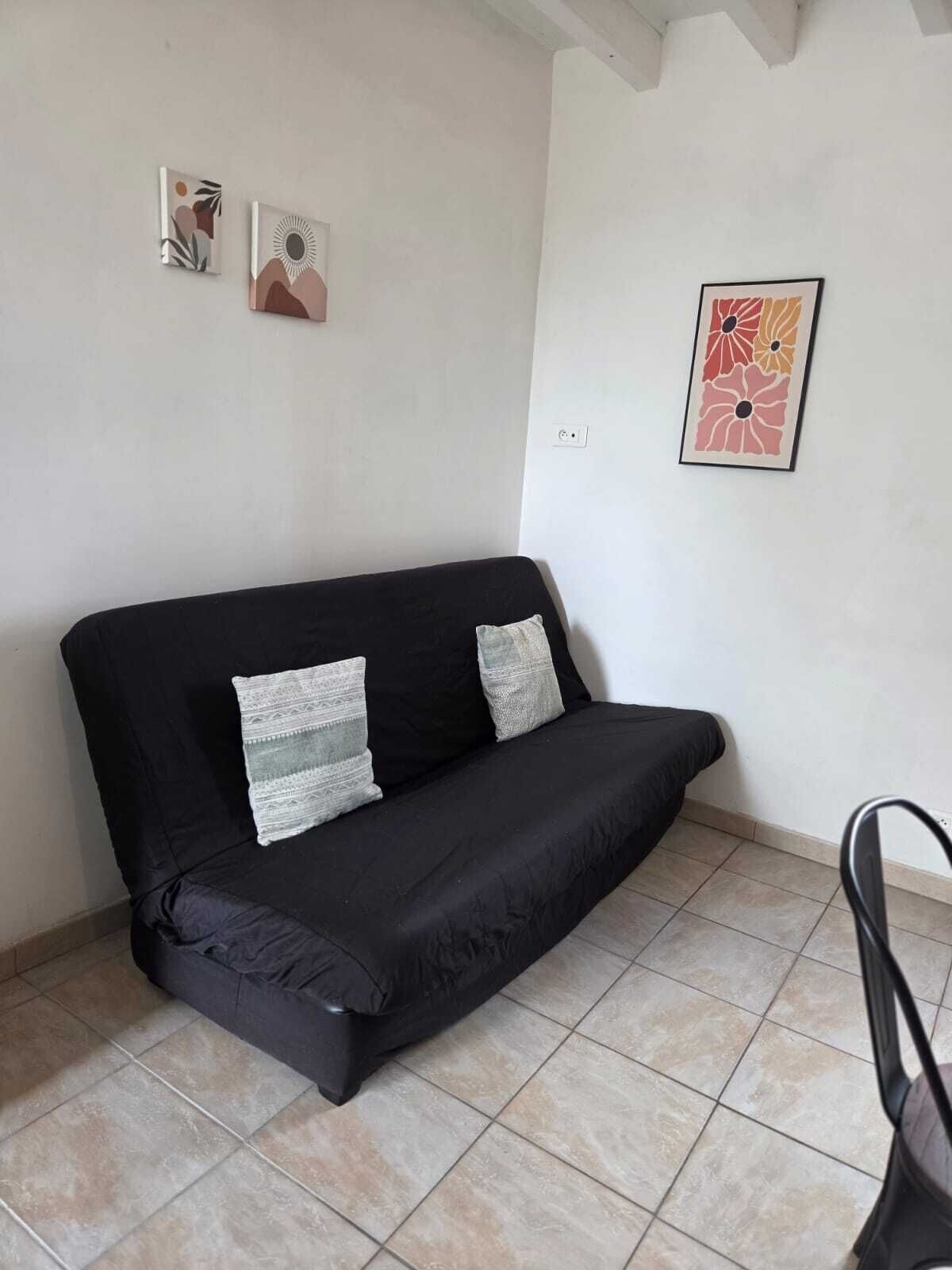Eine schwarze Couch mit zwei Kissen, ein Fliesenboden und zwei gerahmte Kunstwerke an der Wand.