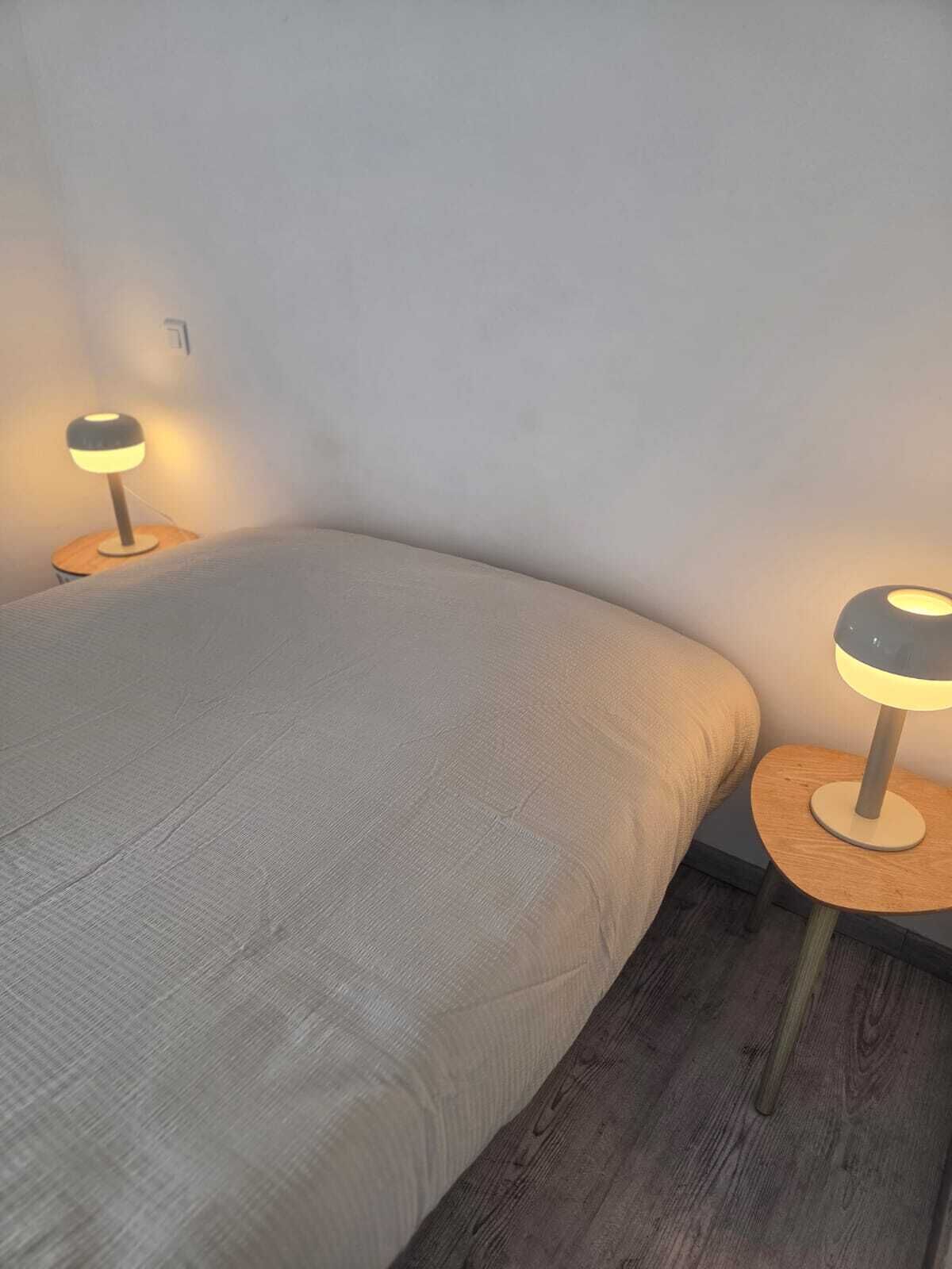 A single bed with a grey blanket in a room with wooden flooring. There are two bedside tables with lamps on either side of the bed.