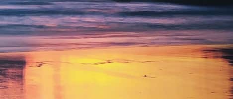 A beach with calm waters reflecting the sunset, distant mountains, and a clear sky.