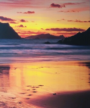 A beach with calm waters reflecting the sunset, distant mountains, and a clear sky.