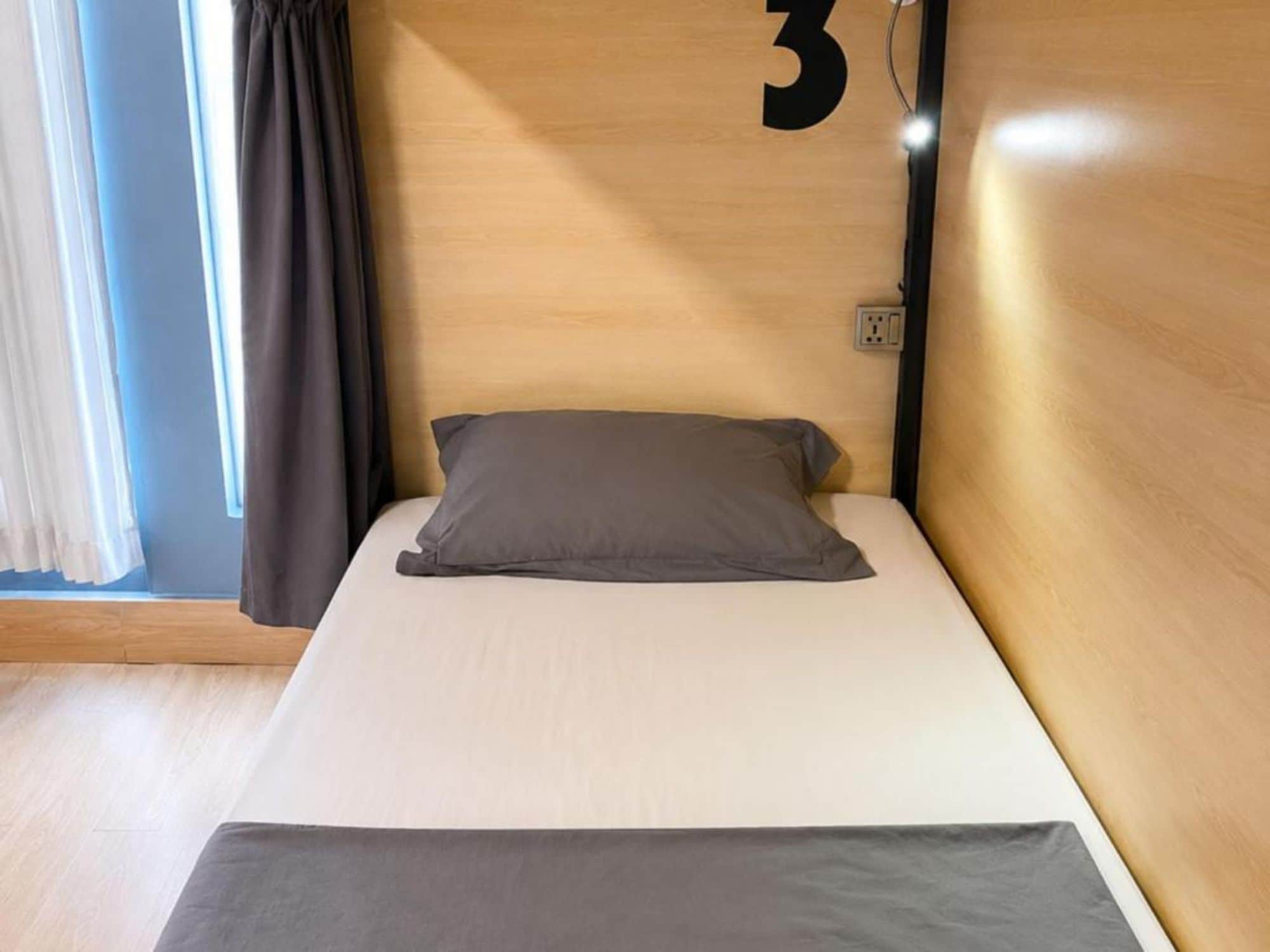 Ein Einzelbett mit grauer Matratze und Kissen, ein hölzernes Kopfende mit der Zahl '3' und eine Steckdose an der Wand.