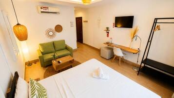 Habitación de hotel con cama, sofá verde, escritorio con silla, televisor y lámpara colgante.