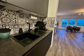 A modern kitchen with a black sink and a coffee machine, a patterned backsplash, and a dining area with a table and chairs. - LEEV Wo 12, Schillig Hörn. 2, Schillig (Schillig)