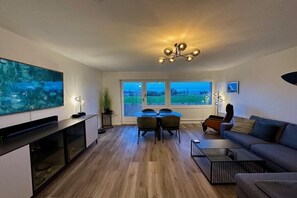 Een moderne woonkamer met een flatscreen televisie, een eettafel met stoelen, een bank en een salontafel. Er zijn ramen met uitzicht op een groen landschap.