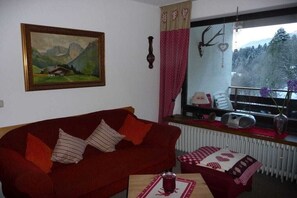 Een woonkamer met een rode bank, een lijst met een landschapsfoto, een radiator en een raam met uitzicht op bomen en bergen.