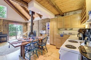 Interior de cabana rústica amb zona de menjador, cuina i finestra gran amb vista a vegetació.