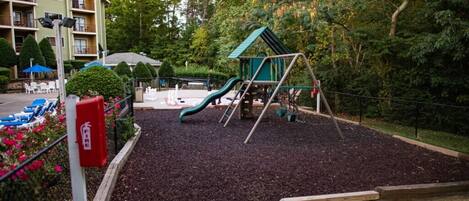 Un parque infantil con un tobogán y estructuras para escalar, rodeado de un área ajardinada con acolchado y un hidrante.