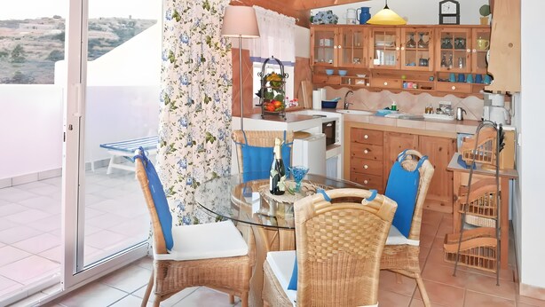 Een eethoek met rieten stoelen en een glazen tafel, een keuken met houten kastjes en een spoelbak, en uitzicht op het landschap door een glazen deur.