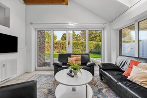 A modern living room with a black leather sofa, a white coffee table, and a black leather armchair. There is a flat-screen TV mounted on the wall, and a large sliding glass door leading to a garden. - Modern beachfront apartment with exceptional sea views at Schönberger Strand (Schönberg (Holstein))