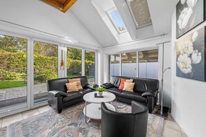A spacious living room with a black leather sofa, a round coffee table, and a large window offering a view of a garden. - Modern beachfront apartment with exceptional sea views at Schönberger Strand (Schönberg (Holstein))