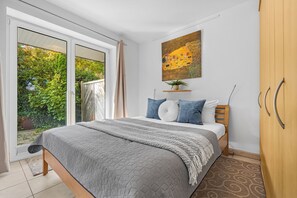 A bedroom with a bed, a sliding glass door to a garden, a painting on the wall, and a wooden wardrobe. - Modern beachfront apartment with exceptional sea views at Schönberger Strand (Schönberg (Holstein))