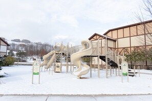 Ein Spielplatz mit Rutschen und Klettergerüsten, umgeben von einem Gebäude mit einer braunen Fassade und verschneitem Boden.