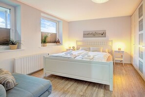 A bedroom with a white bed, bedside tables, a window with blinds, a radiator, and a wall-mounted picture. - Holiday apartment with 1 bedroom (St. Peter-Ording)