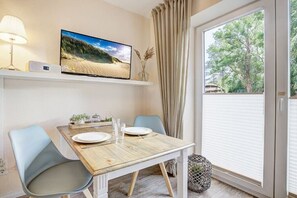 Un comedor con una mesa de madera, dos sillas, un televisor en la pared y vista a árboles por la ventana.