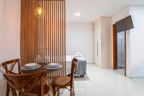 A modern room with a dining table set for two, a bed with a blue cover, and a TV mounted on the wall. - Modern in Cond. w/ Parking at Rio Tavares Ala0004 (Florianópolis)