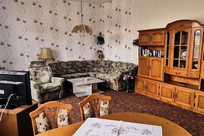 A living room with a sectional sofa, a wooden cabinet, a television, and a dining table. - Holiday apartment Assenta (Blankenburg)
