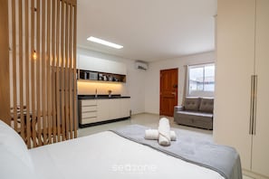 Ein modernes Schlafzimmer mit einem Bett, einer Holztrennwand, einer Küchenzeile, einem grauen Sofa und einem Fenster.