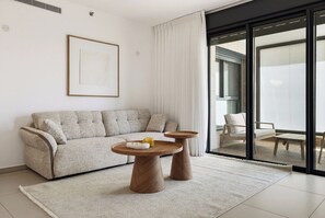 A modern living room with a sofa, two wooden side tables, a rug, and a large window with curtains. - Cozy & Spacious 3BR with Balcony & Mamad (Bat Yam)