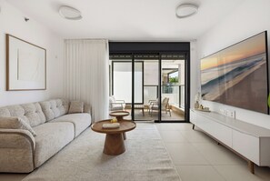 A modern living room with a sofa, a coffee table, a TV, and a sliding glass door leading to a balcony. - Cozy & Spacious 3BR with Balcony & Mamad (Bat Yam)