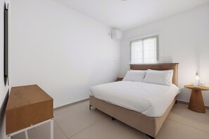 A minimalist bedroom with a bed, bedside table, and a small wooden cabinet. - Cozy & Spacious 3BR with Balcony & Mamad (Bat Yam)