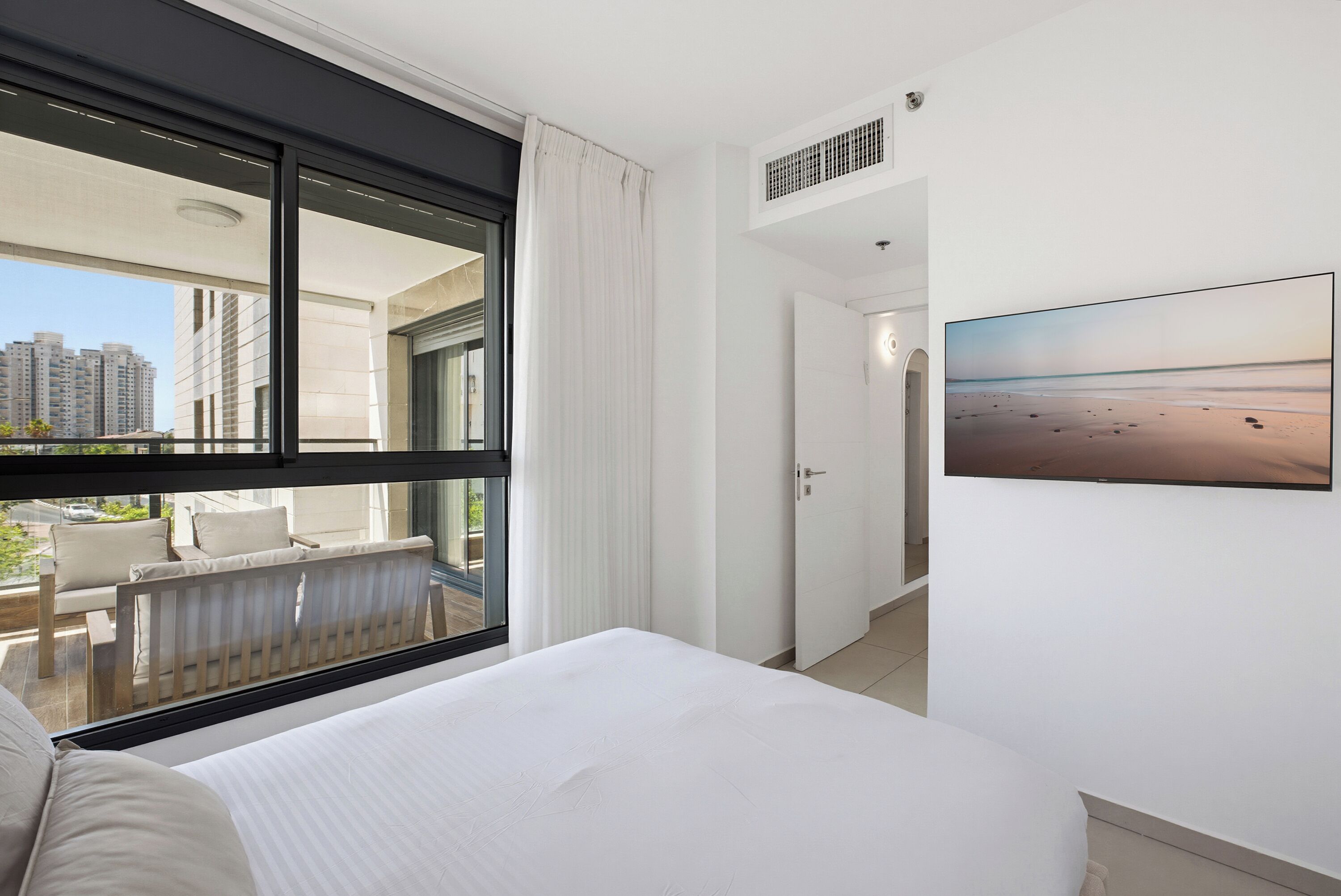 Habitación de hotel con cama, ventana con vista a edificios, una puerta y un cuadro colgado en la pared.