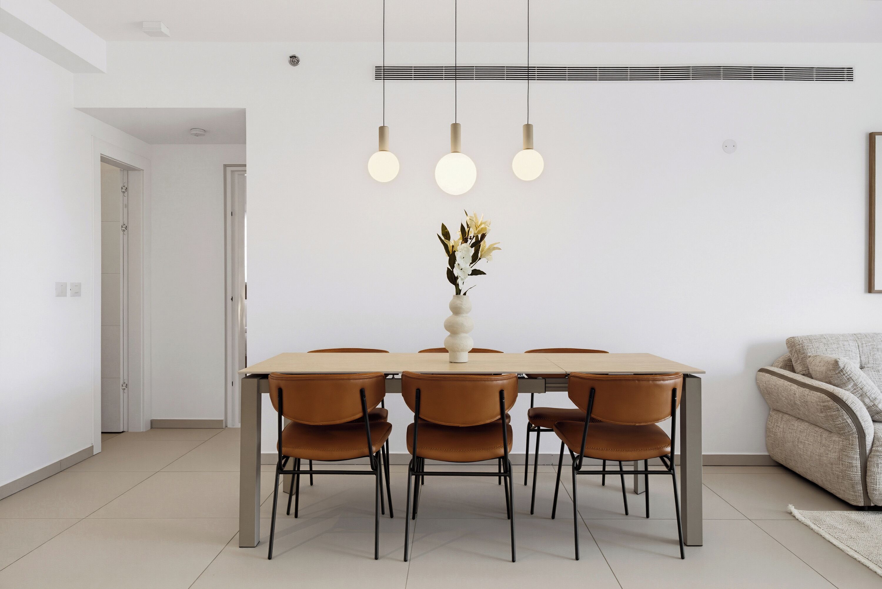 Un comedor con una mesa rectangular, cuatro sillas, un florero con flores y luces colgantes.