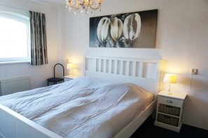 A bedroom with a bed, a nightstand with a lamp, a wall-mounted painting, and a window with curtains. - Residence Juliana 45 (Julianadorp)