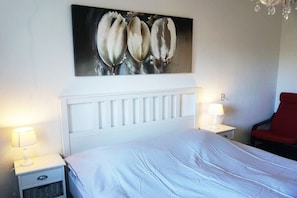 Ein Hotelzimmer mit einem weißen Bett, zwei Nachttischen mit Lampen, einem roten Sessel und einem Wandbild.